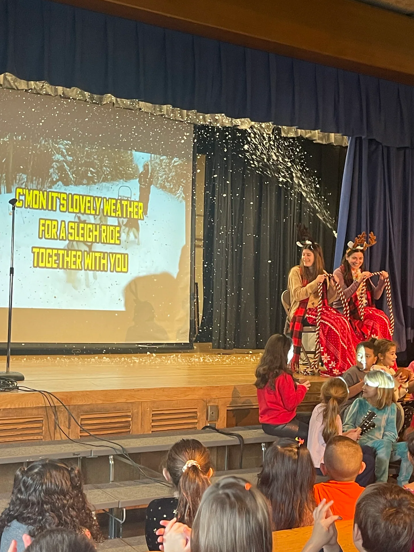 Elementary teachers dancing in a holiday-themed school assembly with indoor snowfall and live music